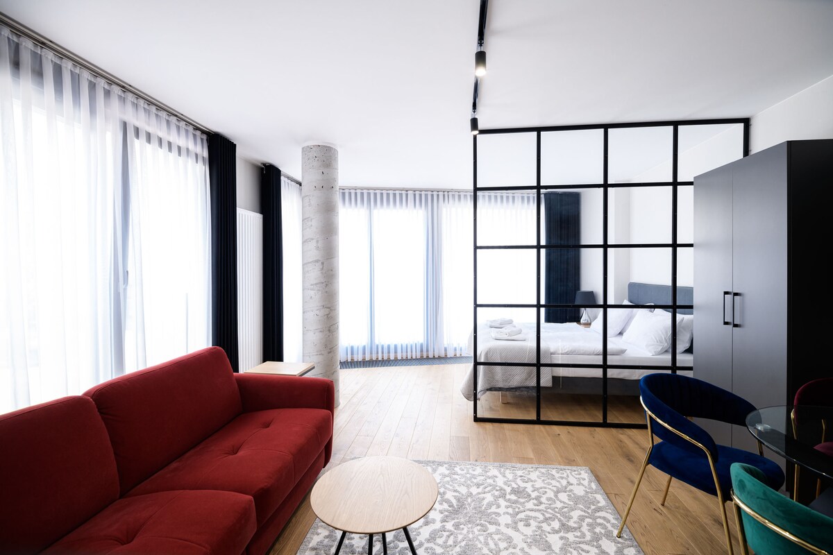A modern living space features a red sofa, a round coffee table, and a glass partition separating the sleeping area. Large windows allow natural light to fill the room, complemented by light curtains. A stylish black wardrobe is positioned nearby.