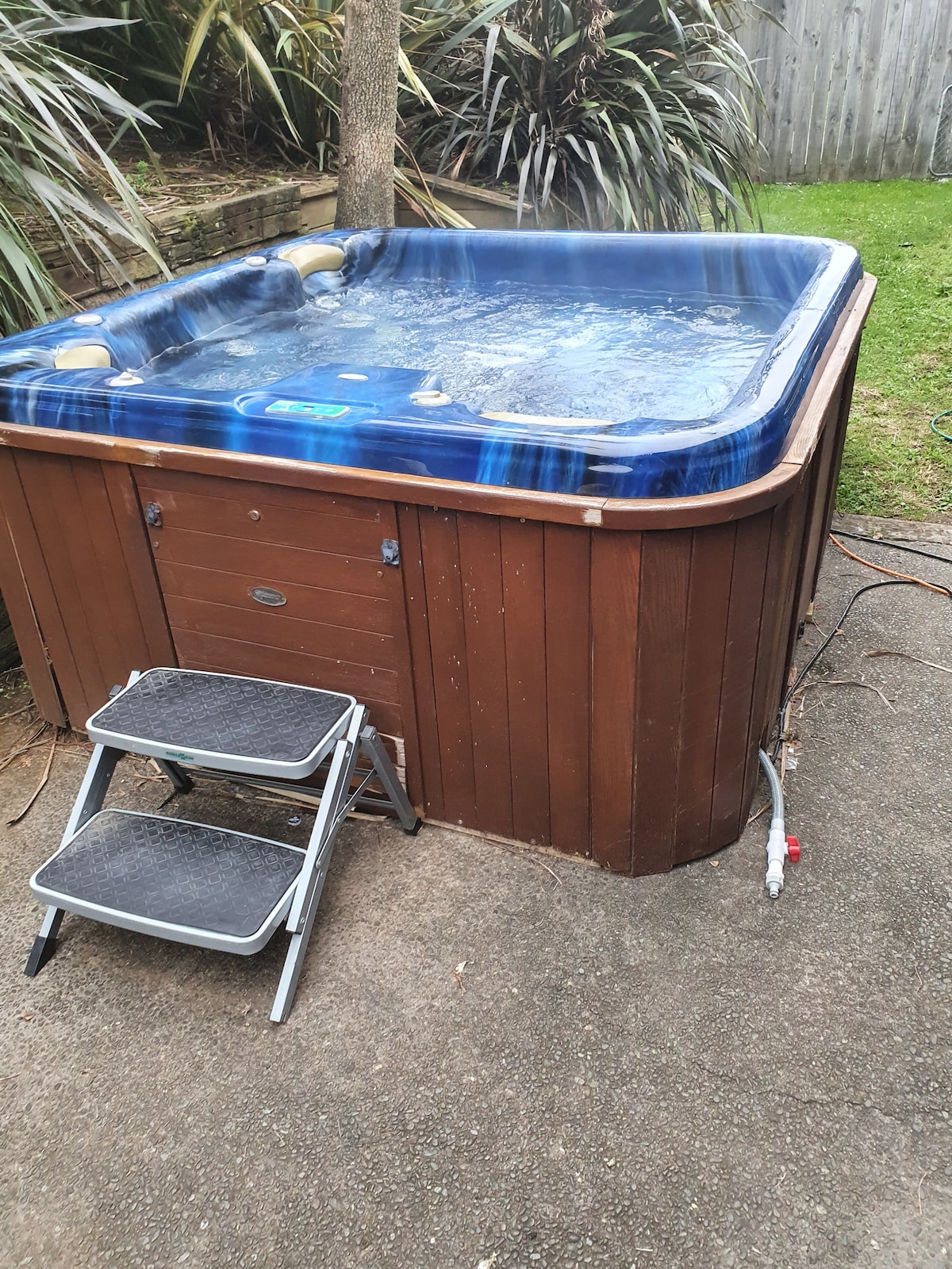 A well-maintained hot tub is set outdoors, surrounded by natural greenery. The wooden paneling complements the blue cover of the tub. Two steps lead up to the hot tub, providing easy access for guests.