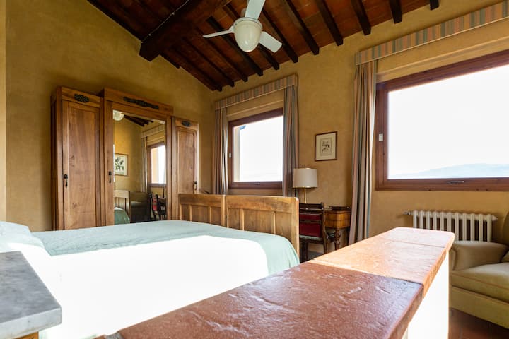 Camera con letto matrimoniale, poltrona, scrittoio, armadio e tre finestre con vista sul paesaggio toscano
