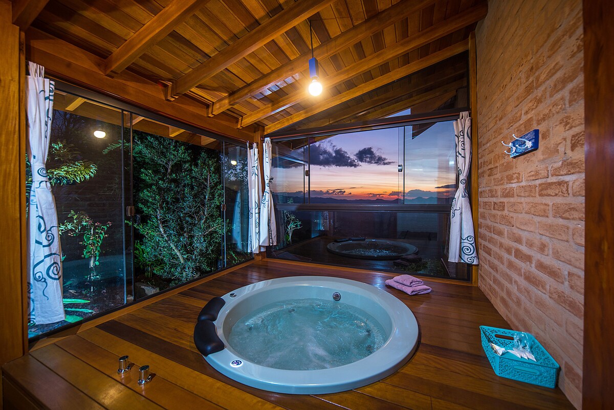 A relaxing indoor area features a circular hot tub with a wooden deck. Large glass windows surround the tub, offering views of the landscape outside. Soft curtains frame the windows, and a small towel lies nearby. The warm wooden ceiling adds a cozy touch to the ambiance.