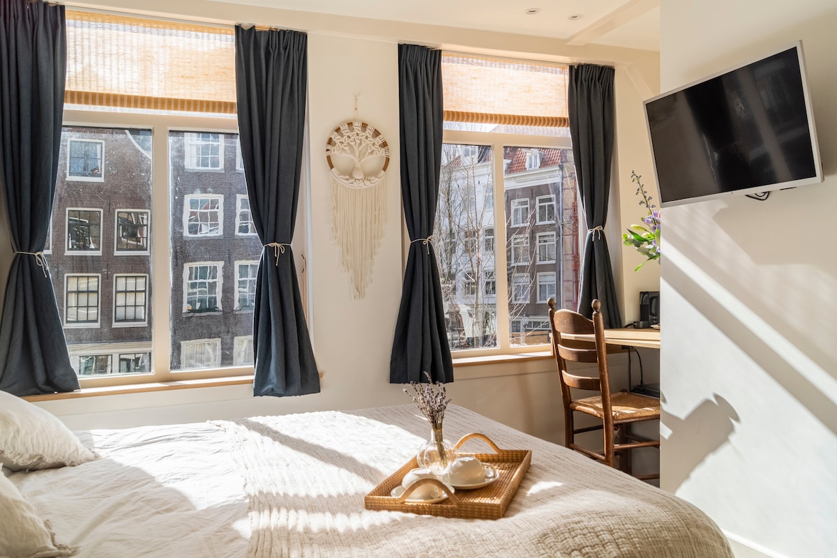 A sunlit room features a spacious bed draped in light linens. Large windows, adorned with navy curtains, provide a view of Amsterdam's historic canal houses. A small desk with a chair sits adjacent to the bed, complemented by decorative elements that enhance the tranquil atmosphere.