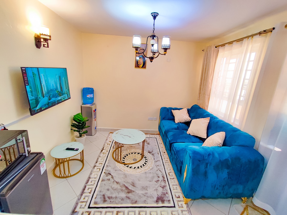 A welcoming living area features a designer blue sofa with decorative cushions, facing a wall-mounted 43" smart TV. Two small round tables complement the space, and a water dispenser adds functionality. Natural light streams in through sheer curtains, illuminating the tiled flooring.