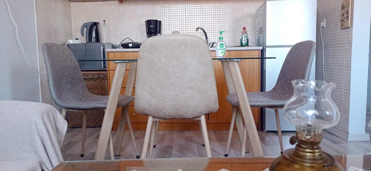 A dining area is presented with a glass-top table surrounded by four muted grey chairs. In the background, a kitchen is visible, equipped with a kettle, coffee maker, and countertop items. A vintage lamp is positioned in the foreground, adding a touch of charm.