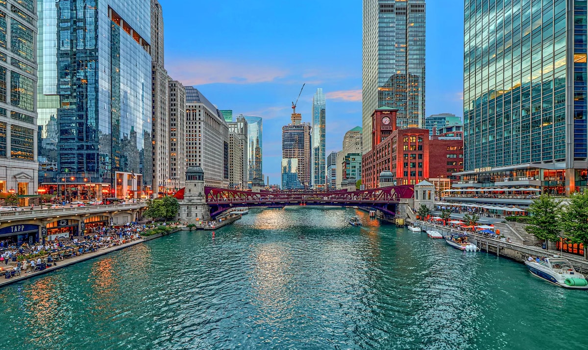 A wide view of the Chicago River, flanked by tall modern buildings. A red bridge crosses the water, with boats visible on the bank. The sky is painted in soft hues as the sun sets, reflecting on the river's surface, creating a serene cityscape.