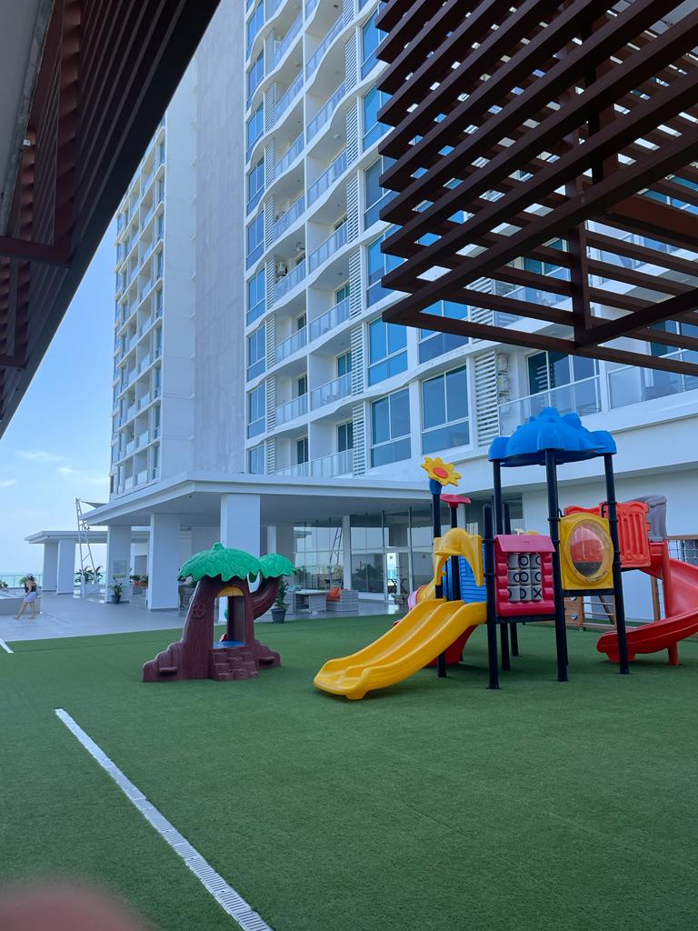 An outdoor play area featuring colorful play structures and slides is situated on green grass. A large building stands in the background, with balconies visible on its facade. The space is well-maintained, providing a welcoming environment for children.