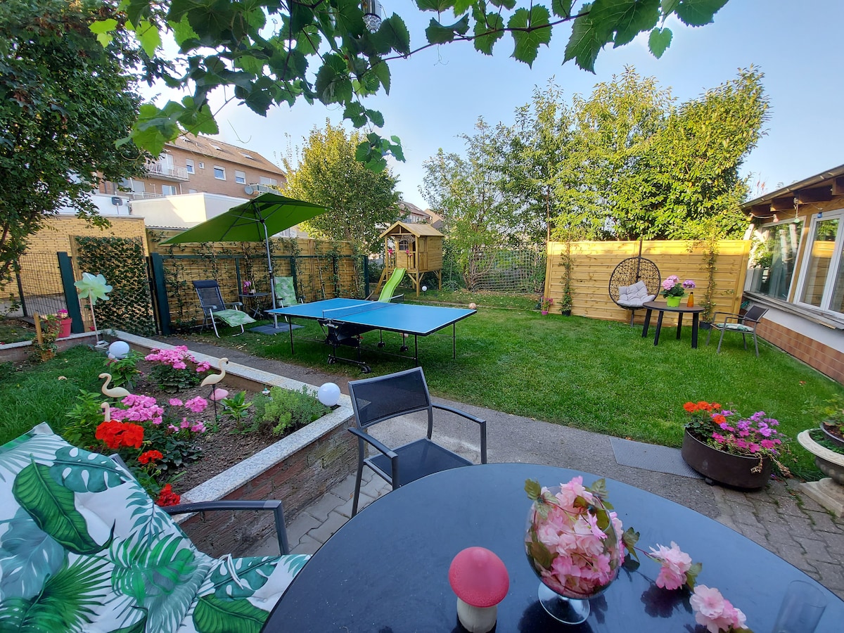 A serene garden space is presented, featuring a well-maintained lawn and vibrant flowers. A table and chairs provide seating options, while a green umbrella offers shade. A ping-pong table is visible, accompanied by a small playhouse, enhancing the outdoor leisure environment.
