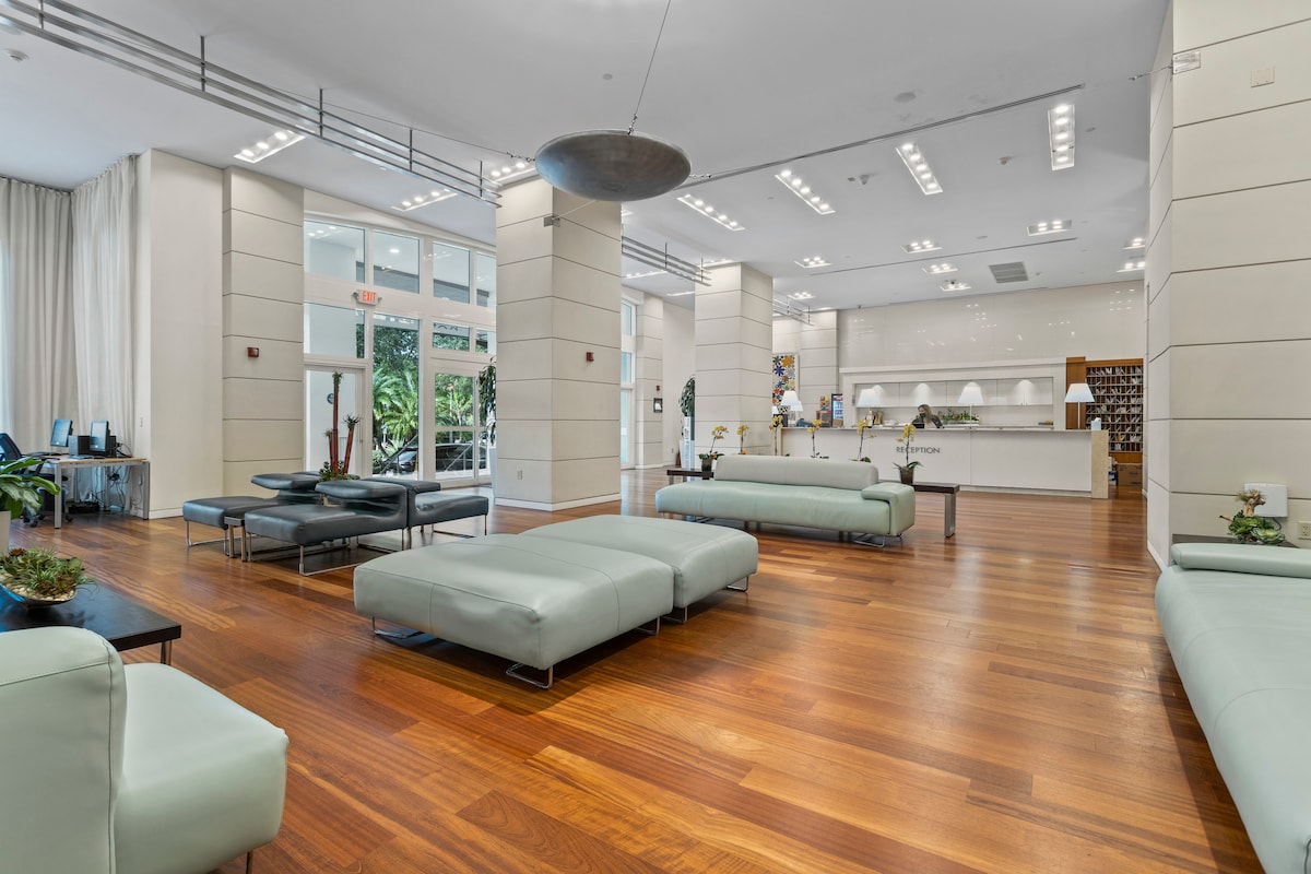 A spacious lobby area features modern furniture with light green seating arranged over polished wooden flooring. Soft lighting enhances the calm atmosphere, while large windows allow natural light to fill the space. A reception desk is visible in the background, providing a welcoming entrance.