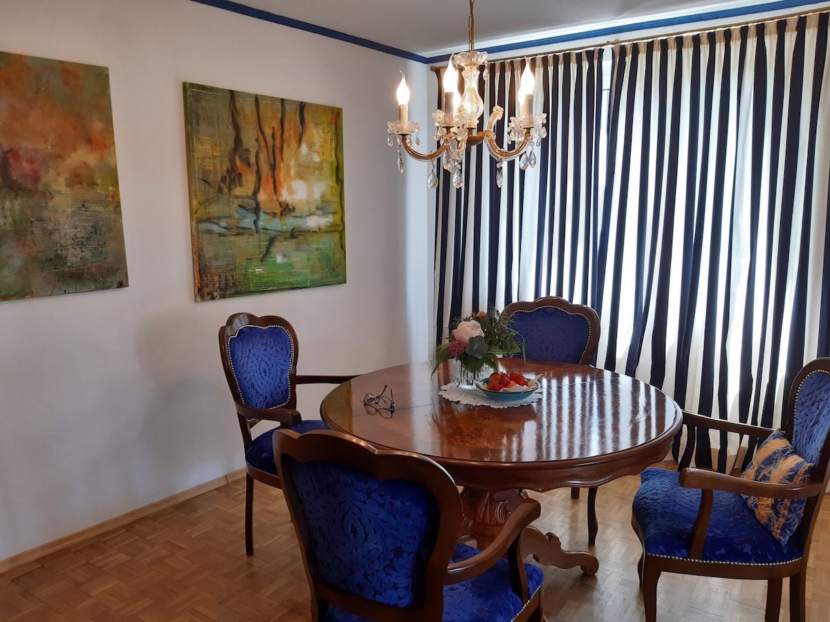 A round wooden dining table is surrounded by four blue upholstered chairs with intricate designs. Two pieces of abstract artwork adorn the walls. A crystal chandelier hangs above the table, and striped curtains allow natural light to filter into the space.
