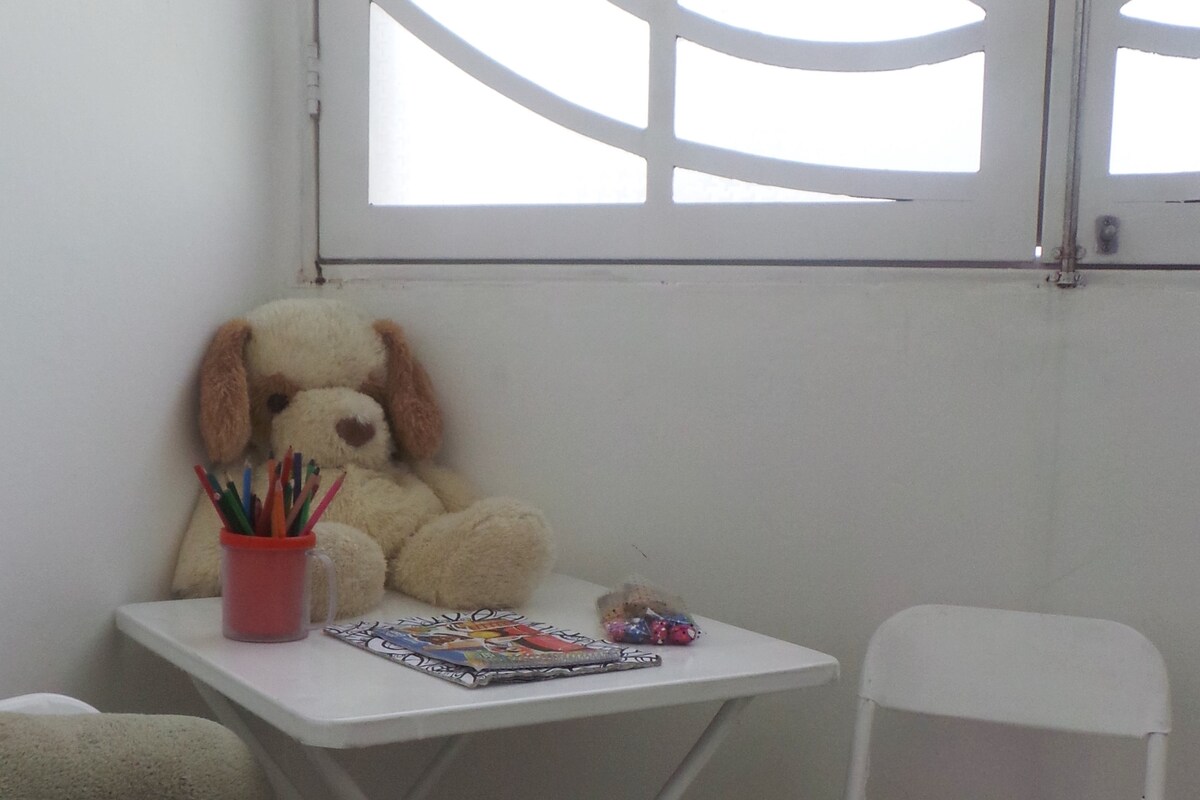 A small table is set against a light-colored wall, featuring a plush dog sitting on a chair. A container filled with colorful pens is placed on the table, alongside a magazine. Soft natural light filters through a window with a decorative design.