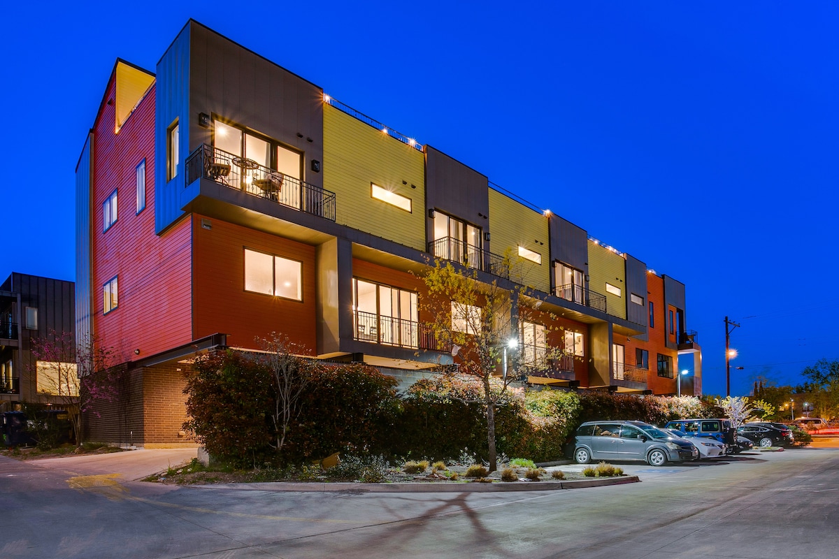 The modern townhouse complex is illuminated against a twilight sky, showcasing a colorful exterior with a mix of red, yellow, and dark hues. Balconies are visible on each floor, and the landscaped driveway includes parking areas beside the building.
