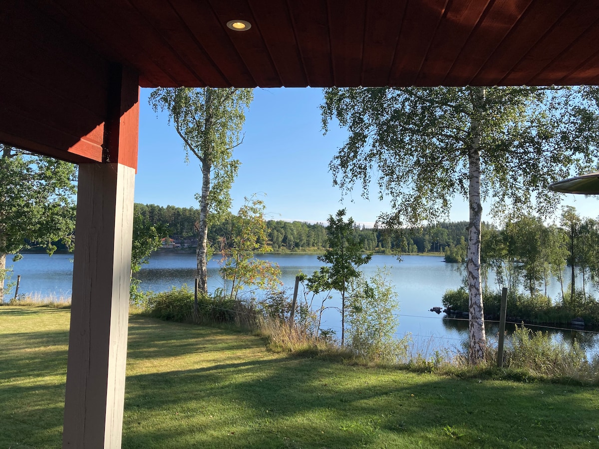 A serene view of the lake is visible from a covered porch. The landscape includes tall trees alongside the shoreline and calm water reflecting the clear blue sky. Lush greenery fills the foreground, providing a tranquil outdoor setting.