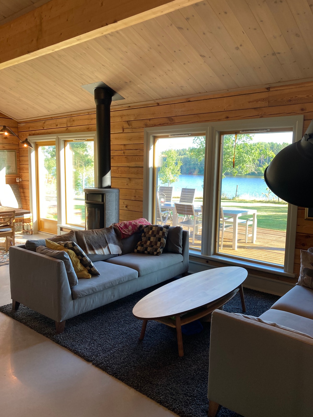 A modern living area is showcased with large windows providing views of the lake. A comfortable sofa is complemented by decorative cushions. A sleek coffee table sits on a textured rug, while a stylish wood-burning stove adds a focal point to the space.