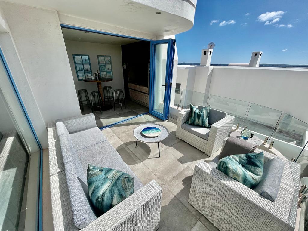An outdoor seating area is positioned on a patio, featuring two cozy chairs and a low table adorned with decorative elements. Large glass doors open to the interior, allowing sunlight to brighten the space while offering stunning views of the surroundings.