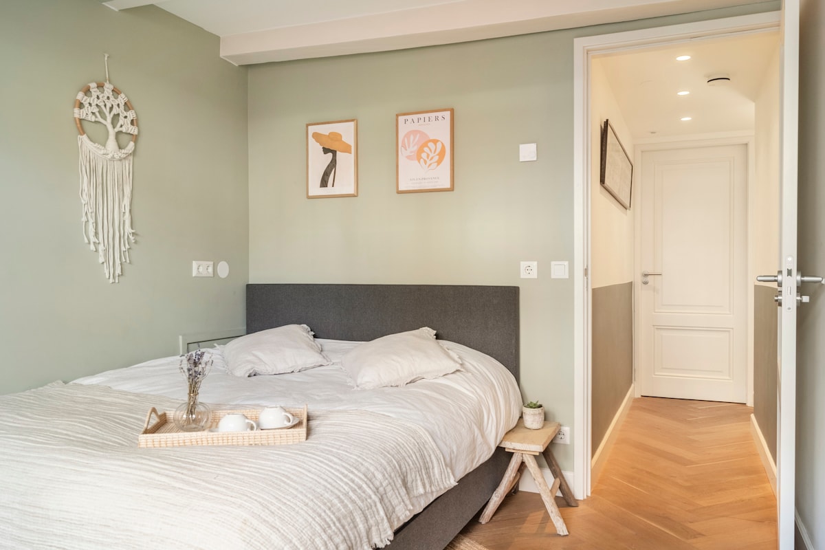 A well-appointed bedroom features a spacious bed with light linens, complemented by two decorative prints on the wall. A bedside table with decorative items sits next to the bed, while a hallway leading to other areas of the house is visible in the background.