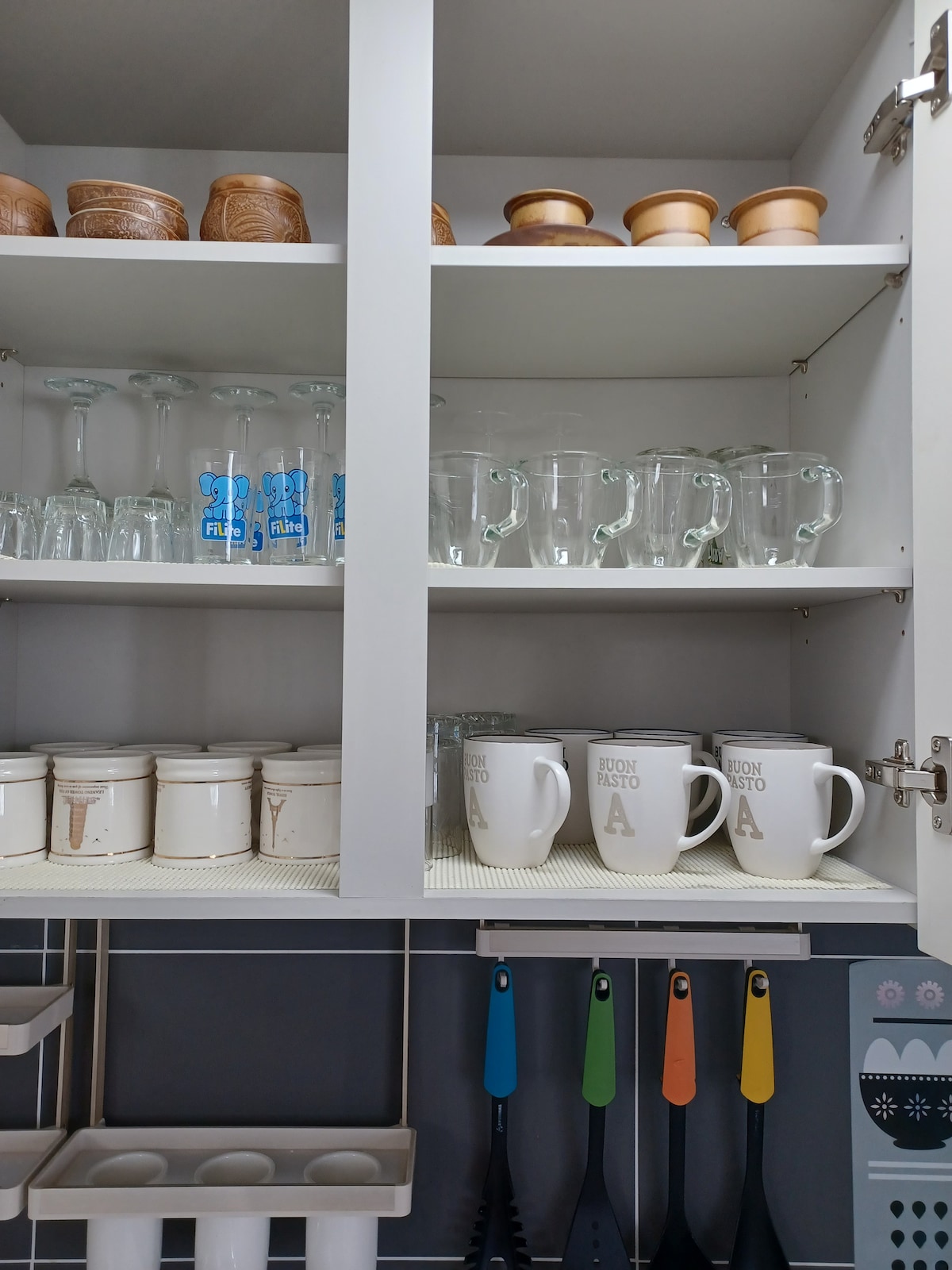 An organized kitchen cabinet displays various glassware and utensils. Clear glass cups and mugs are positioned alongside ceramic jars and distinctively designed containers. Brightly colored measuring spoons are visible, highlighting the kitchen's functionality.
