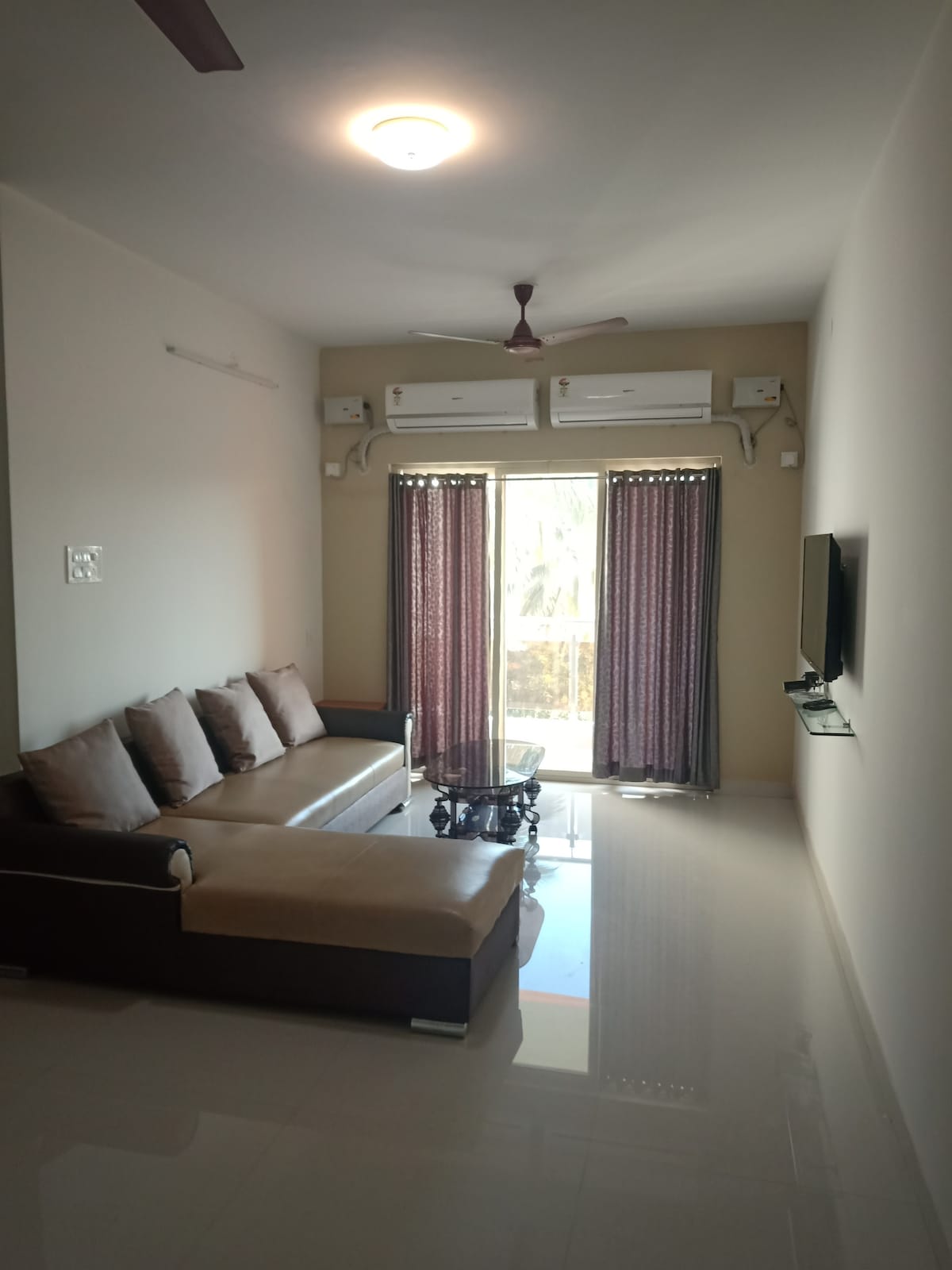 A spacious living room is presented with a comfortable sectional sofa and a glass coffee table. Two large windows with dark curtains allow natural light to filter in, while a wall-mounted TV is positioned for viewing. Air conditioning units are visible above the windows.