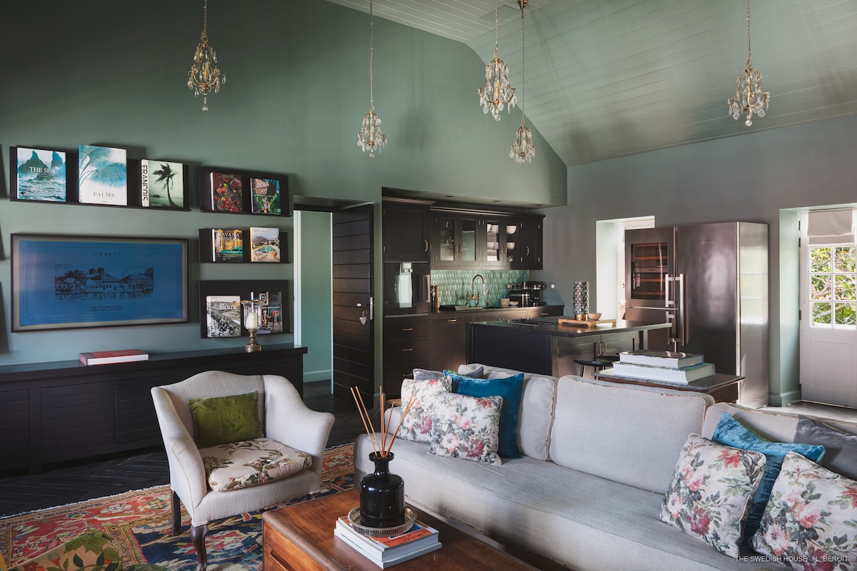 The living room features a spacious seating area with a light sofa and an ornate wooden coffee table. Unique art pieces, including Slim Aarons photography, decorate the walls. A modern kitchen with sleek cabinetry is visible in the background, complemented by elegant crystal chandeliers overhead.