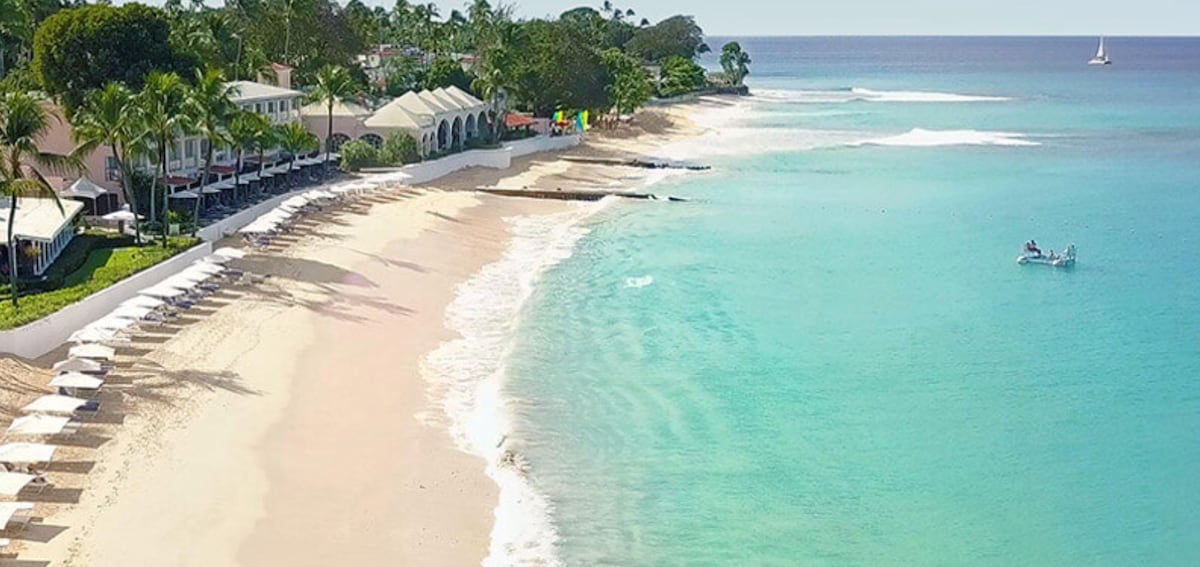 A serene beach stretches along the coastline, featuring soft white sand that meets clear turquoise waters. Beach umbrellas and sun loungers are placed neatly along the shore. In the background, palm trees provide shade, while a sailboat can be seen on the horizon, enhancing the peaceful atmosphere.