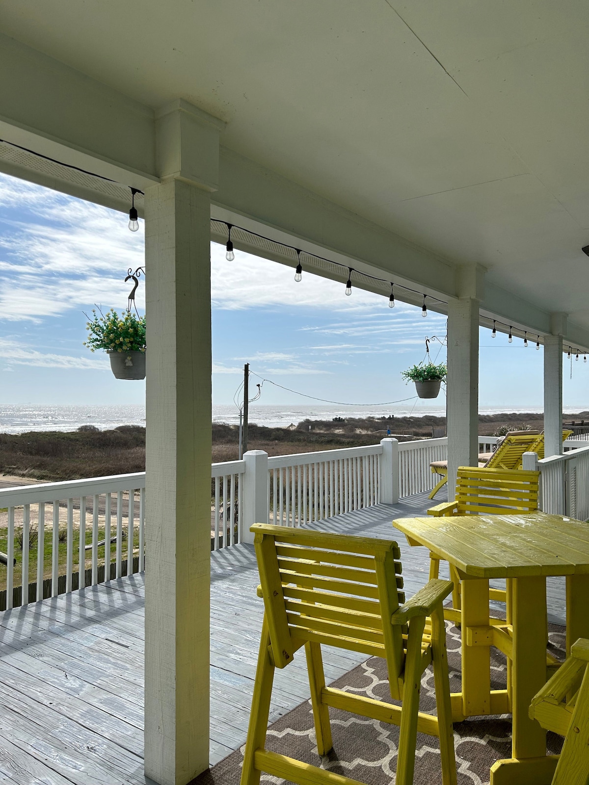 A spacious porch features bright yellow furniture, inviting guests to relax while overlooking the Gulf. Hanging plants add greenery, and string lights provide a welcoming touch. The distant ocean view is visible through the railing, enhancing the outdoor atmosphere.