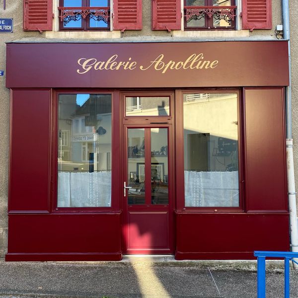 The image displays the front facade of a gallery characterized by a rich burgundy color. Large windows allow natural light to enter, partially covered by white curtains. A glass door invites entry, adding to the inviting appearance of the space.