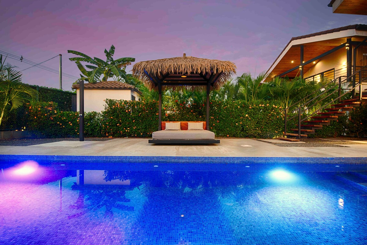 A serene pool area is highlighted by vibrant blue water, complemented by softly lit features. A cabana with a thatched roof and a cushion-covered daybed sits nearby, surrounded by lush greenery and decorative plants, providing a peaceful retreat.