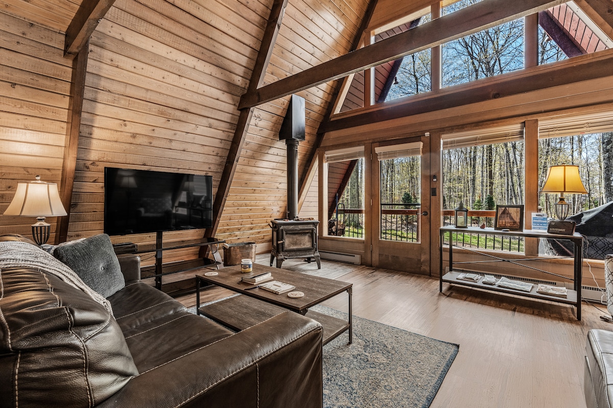A spacious living room showcases a wooden A-frame design with large windows overlooking the woods. Comfortable seating includes a sofa, love seat, and cozy chair arranged around a coffee table. A television is mounted on the wall, and a wood-burning stove adds warmth to the space.