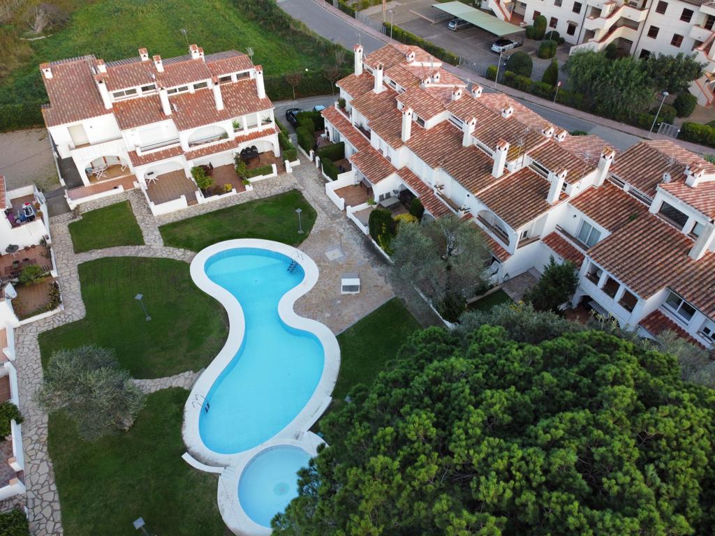 An aerial view captures the community layout featuring a pool with a unique shape surrounded by landscaped gardens. The residential units are visible, showcasing terracotta rooftops and green spaces, while pathways connect various areas of the property.