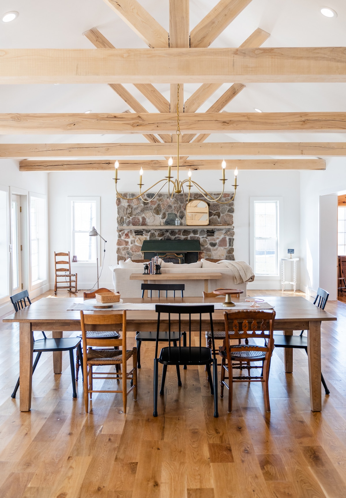 A spacious dining area features a long wooden table surrounded by a mix of black and natural-finish chairs. Exposed wooden beams stretch across the ceiling above, while a stone fireplace adds a rustic element to the room. Large windows allow for ample natural light.