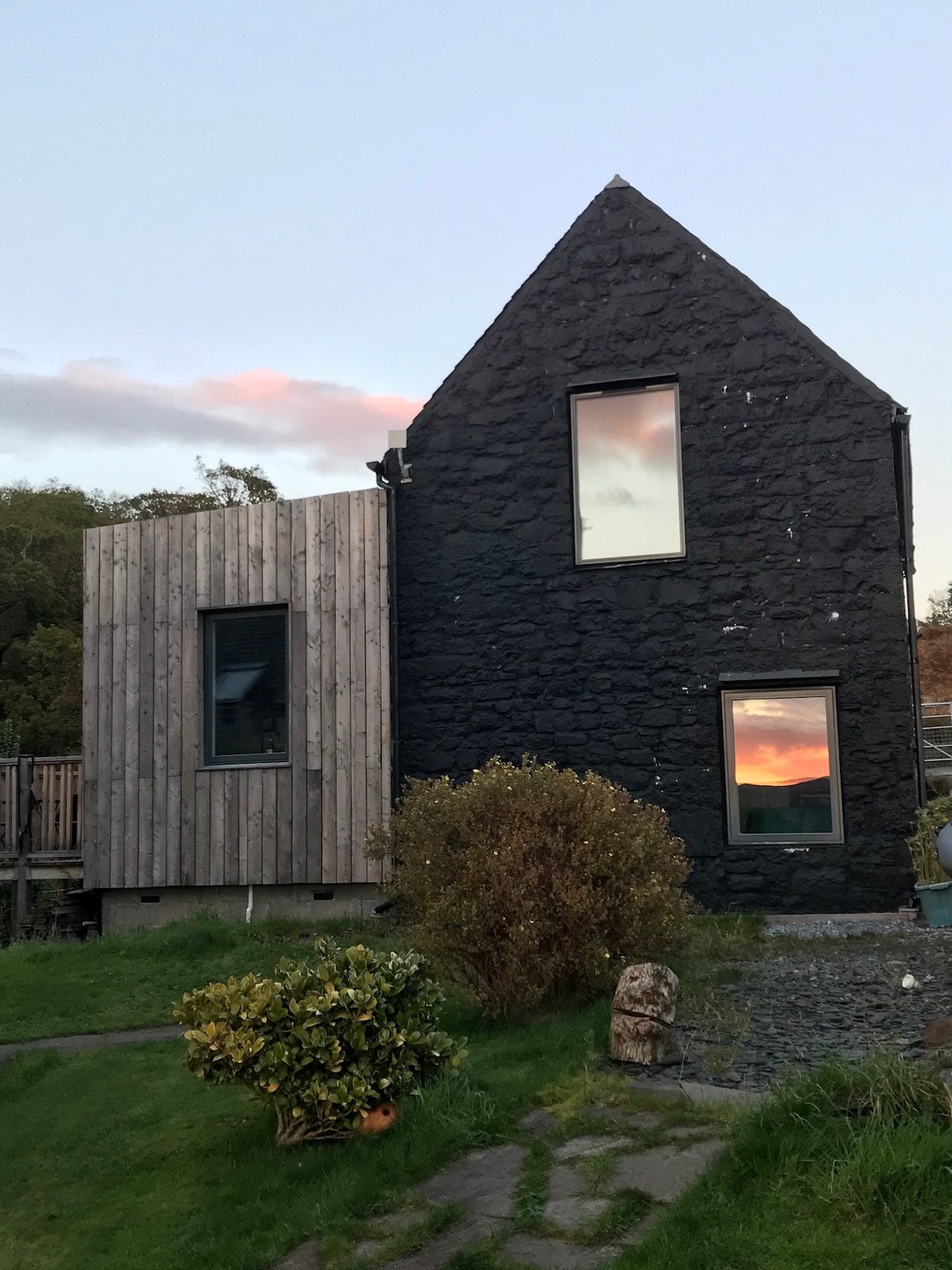 The exterior of the Mill House Steading is displayed, showcasing a contemporary design that blends modern wood and stone materials. The two-tone façade features a dark stone wall alongside a wooden structure, with large windows reflecting the colors of the sunset.