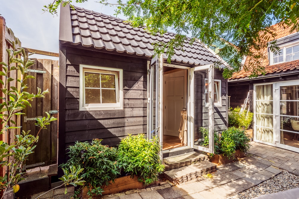 A charming tiny house features a dark wooden exterior with a distinctive tiled roof. Open double doors invite natural light inside, while a stone pathway leads to the entrance. Lush greenery and stone pavers enhance the serene garden setting.