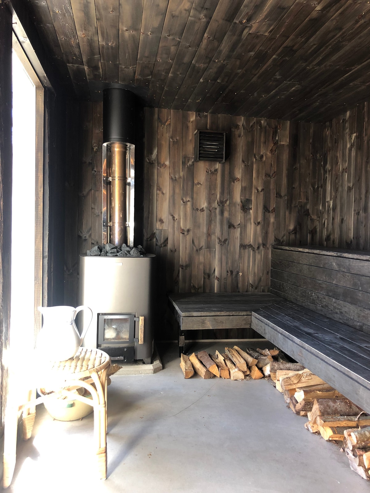 A spacious sauna features wooden walls and benches, creating a warm atmosphere. A stove stands in the corner, accompanied by a stack of firewood nearby. Natural light filters in from a glass door, providing a view of the outdoor surroundings.