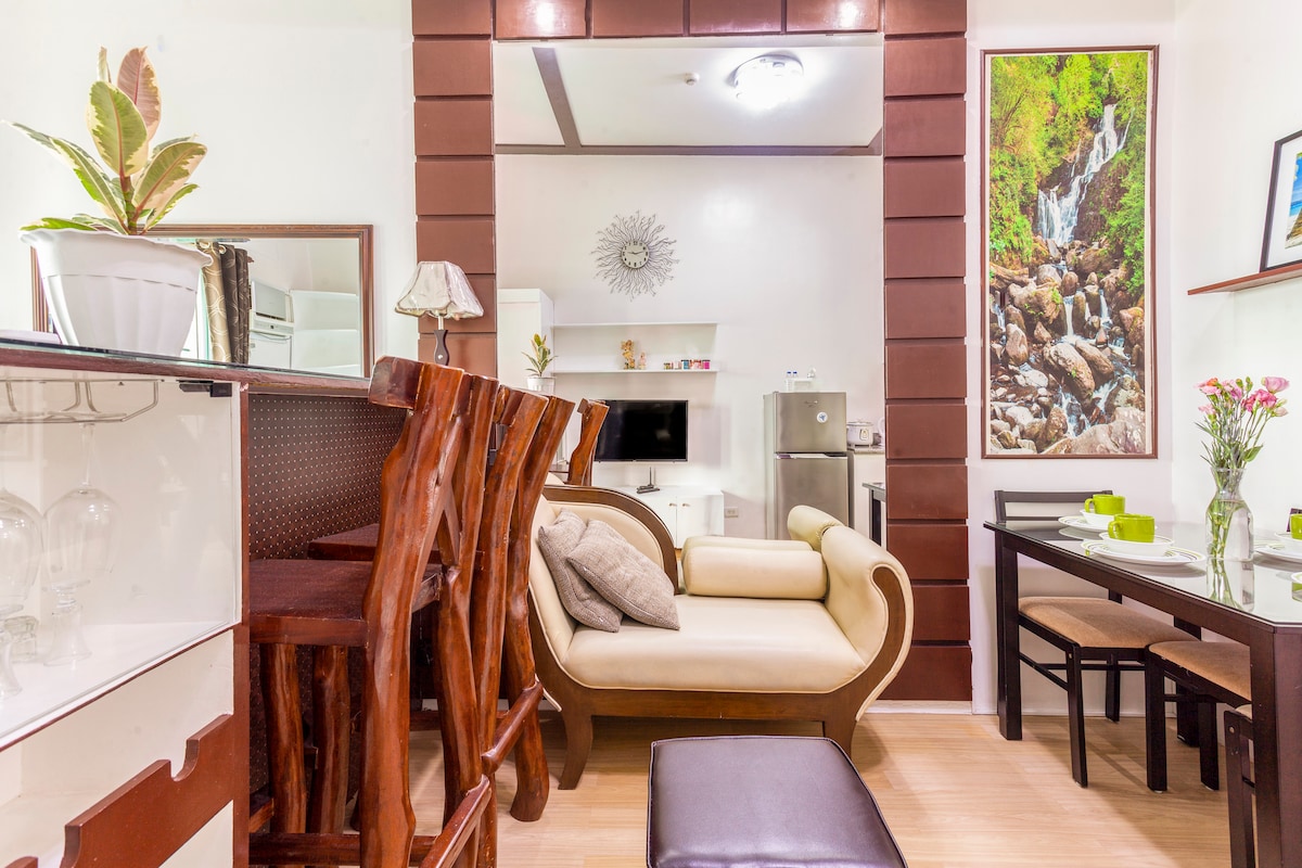 A cozy living area features a light-colored sofa adorned with cushions, complemented by a dark ottoman and a wooden dining table. A decorative wall art piece showcasing a waterfall adds a touch of nature, while modern appliances are subtly integrated into the background.
