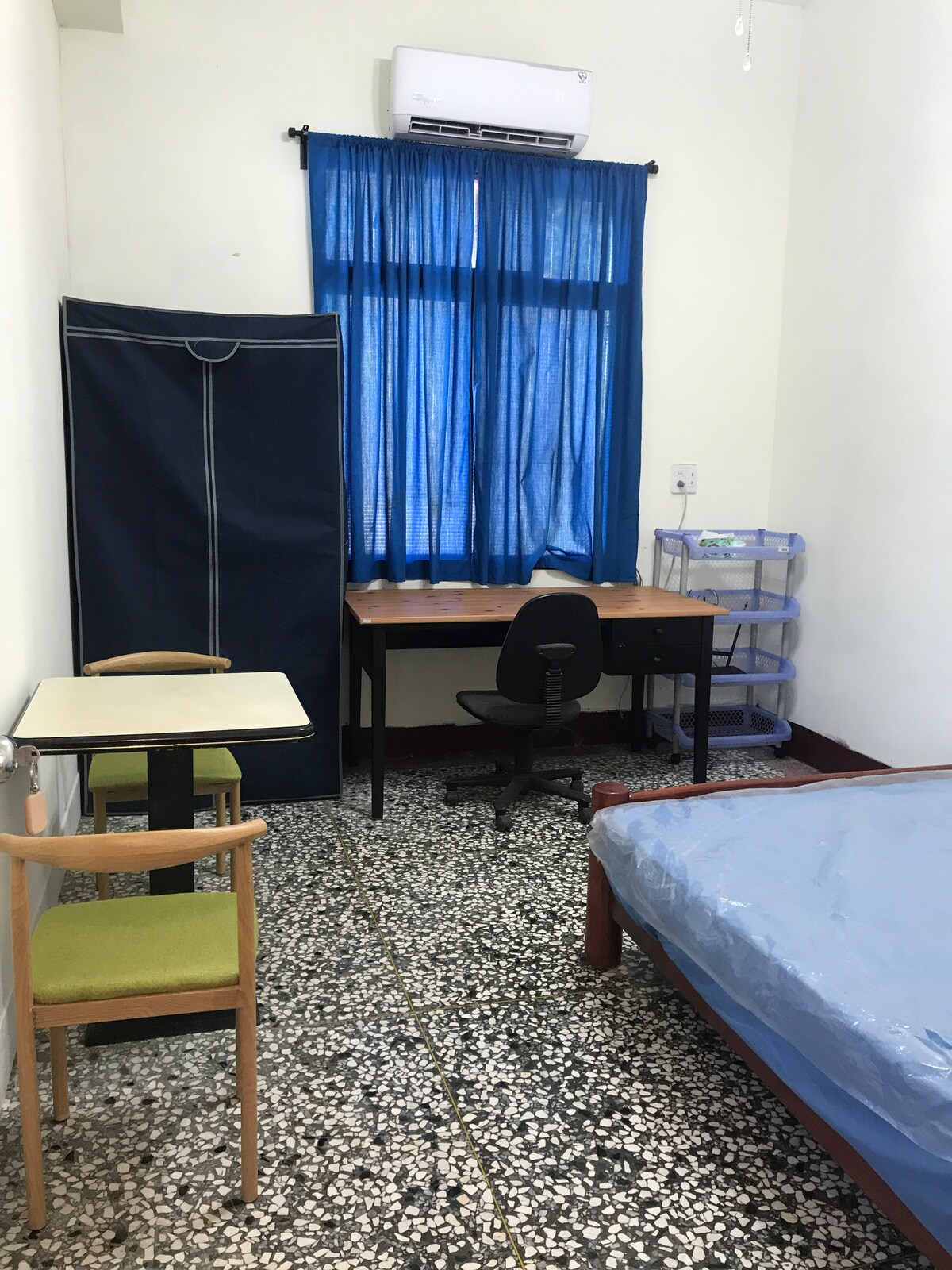 A simple room features a spacious bed with a blue coverlet. A desk with a chair is positioned next to a window dressed in bright blue curtains. A clothing rack stands beside a small table and chair, providing functional elements to the space.
