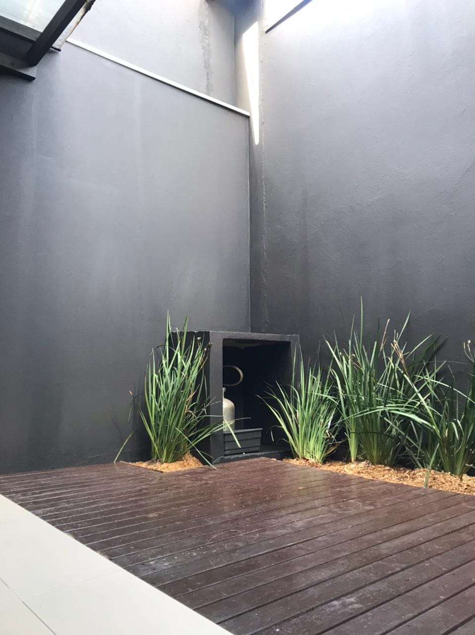 An enclosed outdoor space features dark walls, creating a serene ambiance. A wooden deck runs along the floor, complemented by tall green plants placed on either side. A small alcove is present, adding a focal element to this tranquil area.
