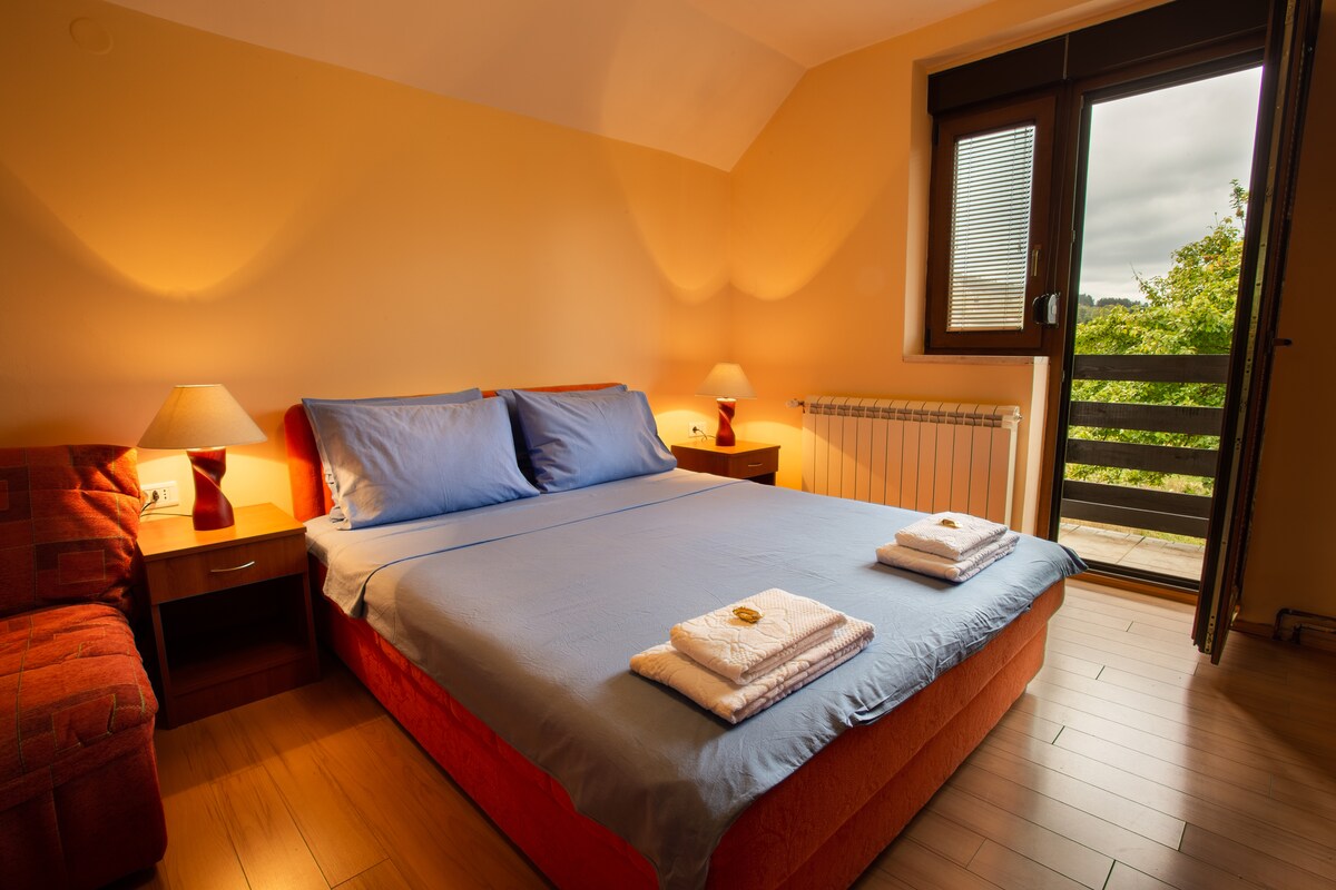 A spacious sleeping area features a plush queen-size bed with soft bedding and neatly arranged towels. Soft lamps provide gentle lighting beside the bed. A large window offers views of lush greenery, while a cozy sofa is placed in the corner of the room.