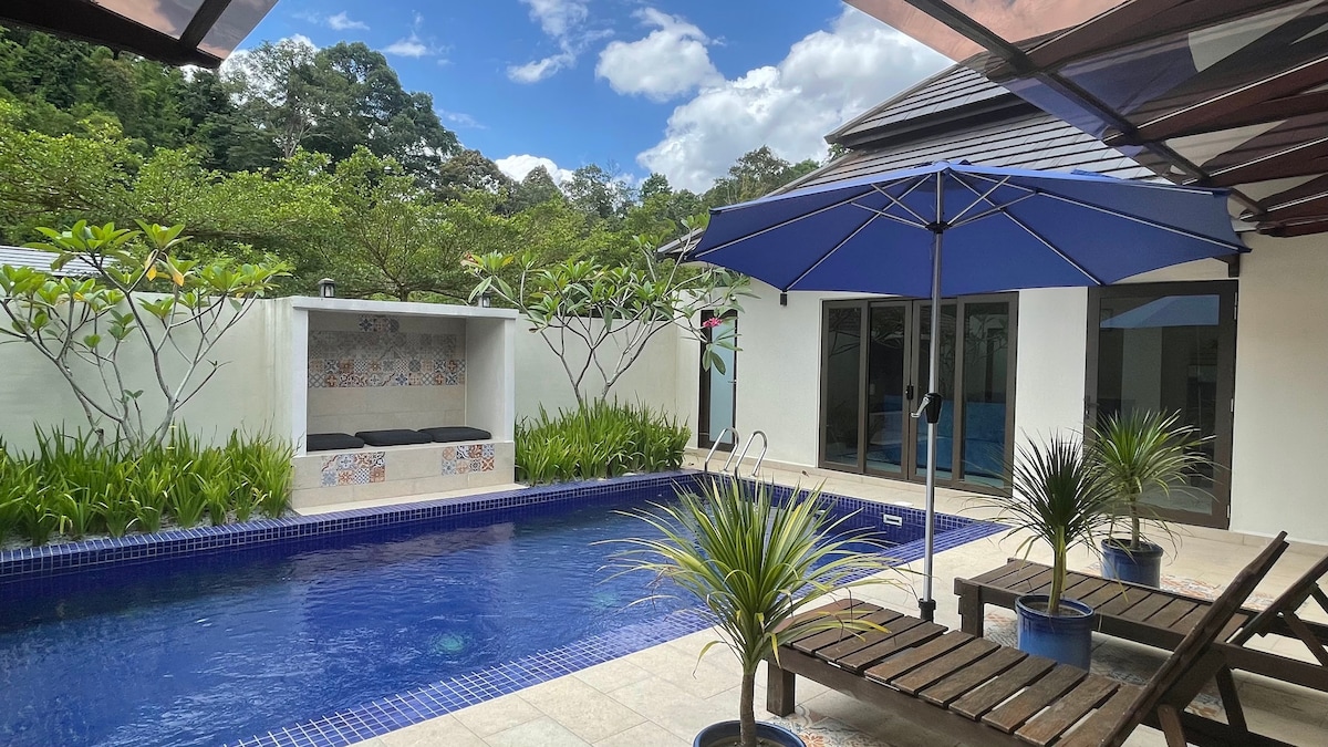 A private pool area is featured, surrounded by greenery and two lounge chairs. An umbrella provides shade over the poolside seating. A small outdoor seating area with decorative tiling and raised plants is visible, creating a tranquil environment for relaxation.