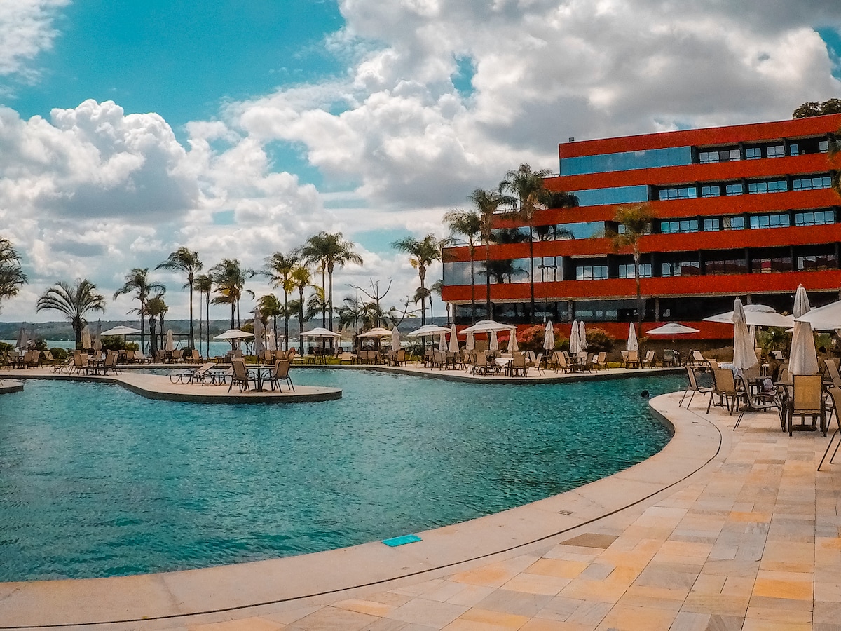 A large, inviting pool is surrounded by lounge chairs and tall palm trees, with umbrellas providing shade. The hotel features a modern red facade, and the scenic landscape blends into a cloud-filled sky, creating a relaxing atmosphere for guests.