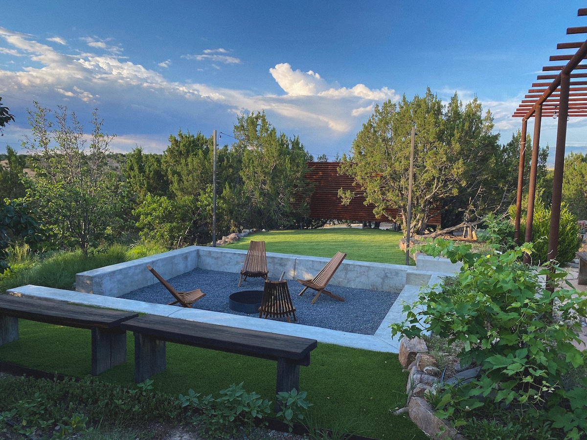 An outdoor communal space features a natural swimming hole surrounded by green grass and trees. Several wooden lounge chairs are positioned around a fire pit on a gravel surface. A wooden bench sits nearby, providing a place to relax and enjoy the tranquil environment.