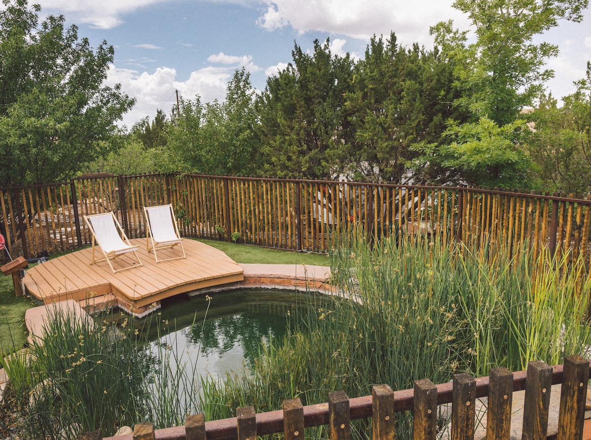 A natural swimming hole is surrounded by lush greenery and tall grasses. Wooden deck loungers are positioned on a platform that leads into the water, while a rustic wooden fence encloses the serene outdoor space, offering a peaceful atmosphere.