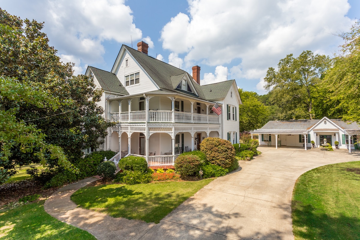 The historic Victorian mansion is framed by lush greenery and features a welcoming wraparound porch with white railings. An American flag flutters from the second floor. A winding driveway leads to a separate building in the background, surrounded by manicured lawns.