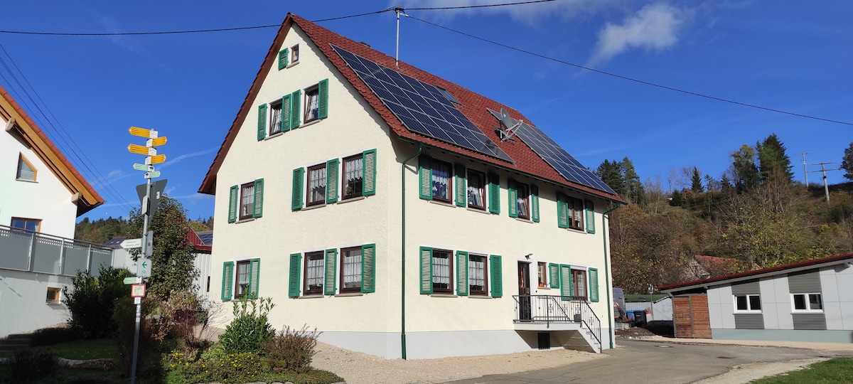 A multi-story building is showcased, featuring a light-colored facade with green shutters and a red roof. Solar panels are installed on the roof, and the entrance is accessible via a small staircase. Surrounding landscaping includes shrubs and a gravel pathway.