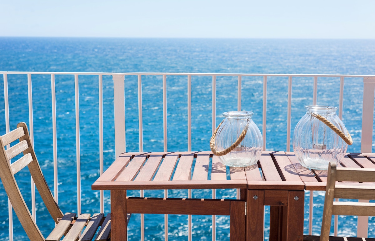 A wooden table with two matching chairs is positioned on a balcony, set against a backdrop of the vast ocean. Two glass jars with handles rest on the table, reflecting sunlight. The seamless blue water stretches into the horizon, emphasizing the serene coastal environment.