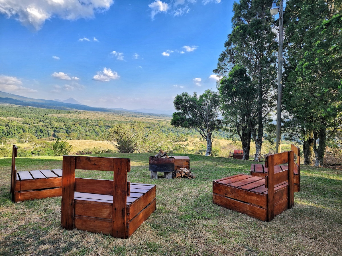 A serene outdoor seating area offers wooden benches arranged around a fire pit, surrounded by vibrant green grass. The expansive view features a valley landscape with distant mountains under a clear blue sky, creating a peaceful setting for relaxation.