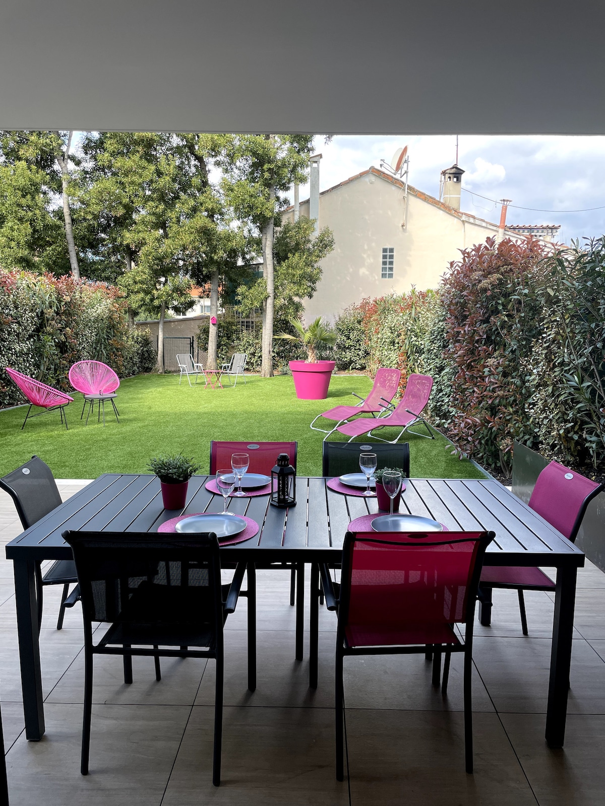 A stylish outdoor dining area features a large table set for six, with vibrant pink plates and black chairs. The private garden is visible in the background, showcasing green lawn space adorned with colorful lounge chairs and decorative plants, creating a relaxing atmosphere.