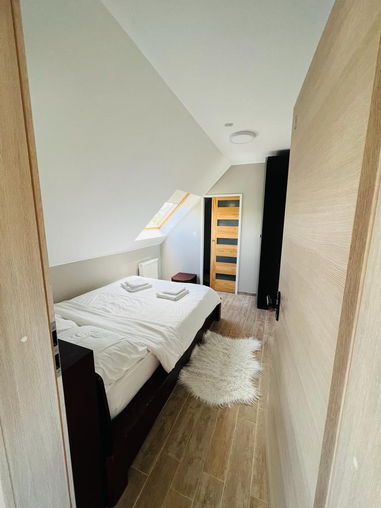 A bright room features a spacious bed with fresh linens and a plush rug positioned on wooden flooring. Natural light filters through a slanted window, and a modern closet stands opposite the entrance, enhancing the room's clean and functional design.
