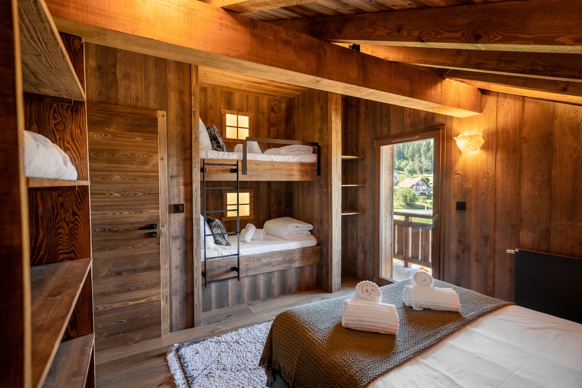 A spacious bedroom features warm wooden walls and beams, with a configuration of a large double bed and a bunk bed in the corner. Natural light filters through the window, revealing a view of the surrounding landscape. Fresh towels are neatly arranged on the beds.