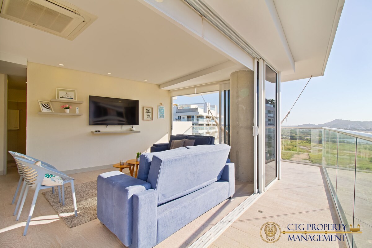A comfortable living area features a blue sofa facing a wall-mounted television. Natural light filters through large glass doors, which open to a wrap-around balcony. A small coffee table is positioned on a light carpet, contributing to the open and airy atmosphere.