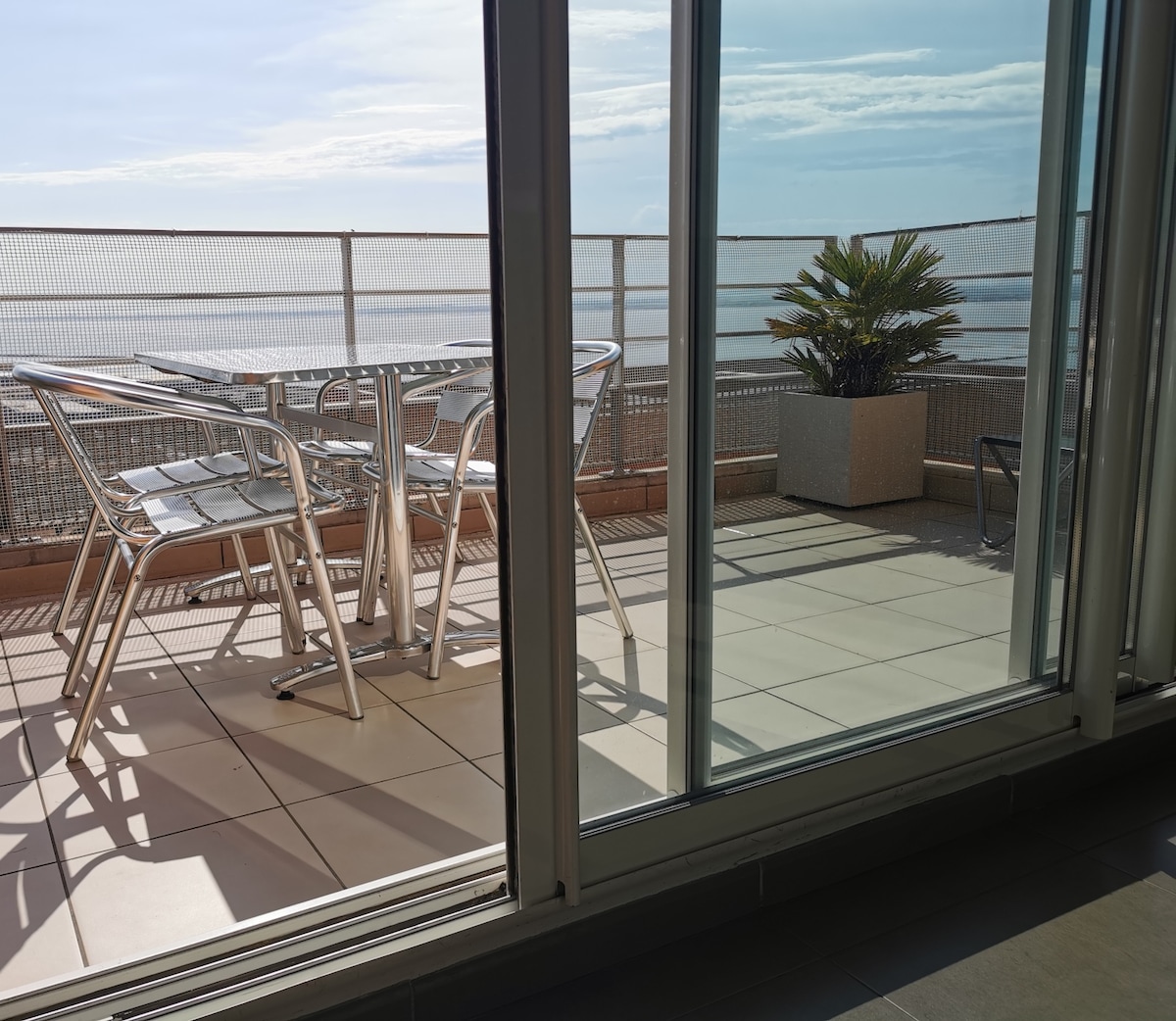 A balcony is visible through large glass doors, featuring a table and chairs, accompanied by a potted plant. The tiled flooring extends outdoors, with a panoramic view of the surrounding landscape beyond the railing.