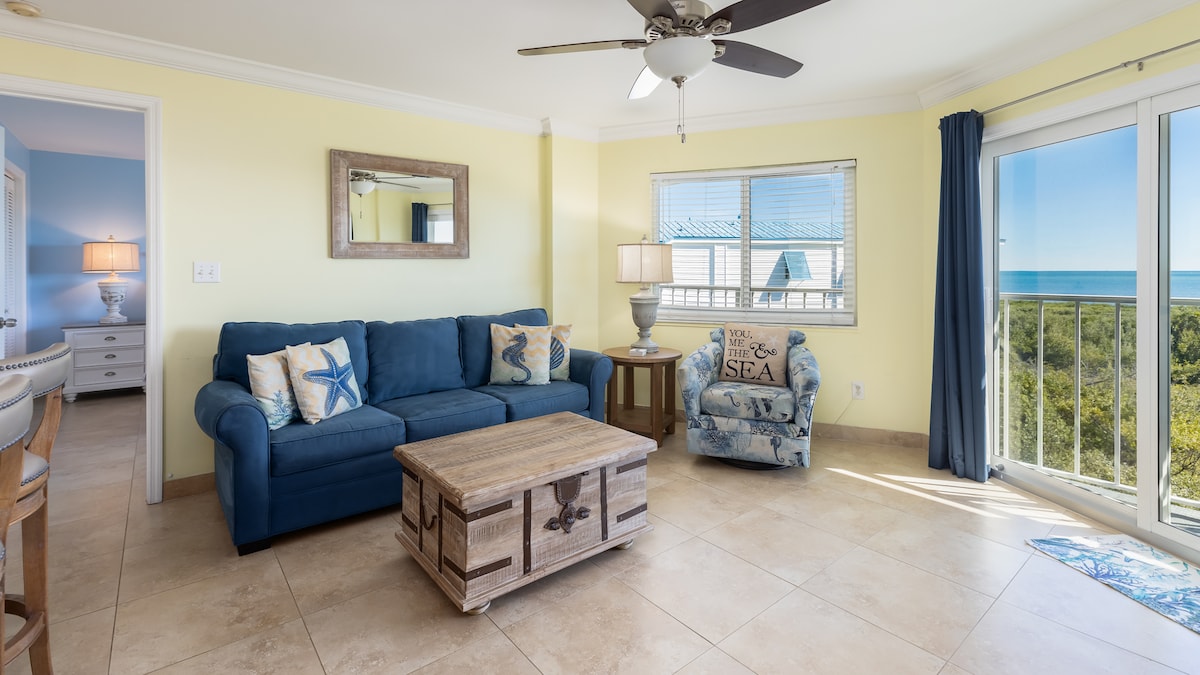 A bright living room features a blue sofa and an accent chair with coastal patterns, complemented by a wooden coffee table. Large windows reveal views of the ocean and mangroves, while a ceiling fan provides a refreshing breeze in this airy space.