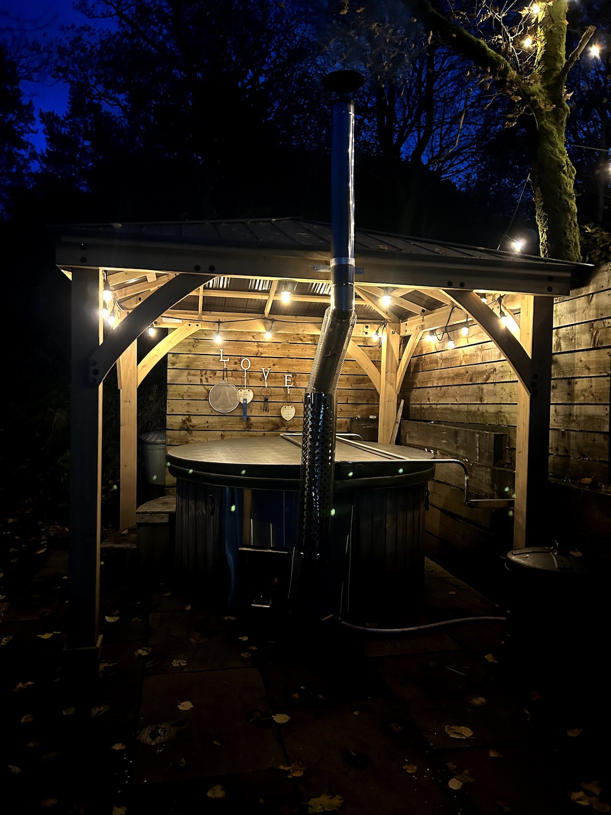 A wooden structure houses a wood-fired hot tub, surrounded by warm string lights that create a cozy ambiance. The beautifully crafted space features wooden walls and a tiled floor, inviting relaxation under the evening sky.